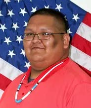 Officer Elmer Lamson stands in front of an American Flag in a red shirt and necklace.